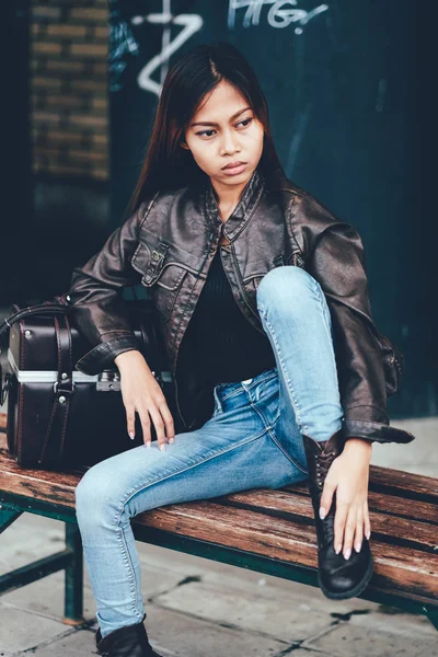 Linda jovem mulher em jaqueta de couro, sentado no banco posando com saco de couro, hipster, estilo Vintage Retro — Fotografia de Stock