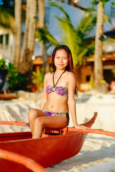 Menina bonita feliz em um maiô de verão sentado em um velho barco de pesca na praia de areia branca tropical. Conceito férias de verão — Fotografia de Stock