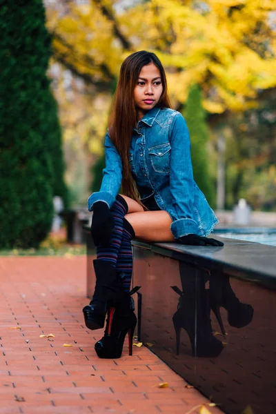 Wanita muda cantik mengenakan jaket jeans biru dengan perbukitan tinggi dan kaus kaki panjang bergaris-garis lutut. Gadis yang duduk di taman — Stok Foto