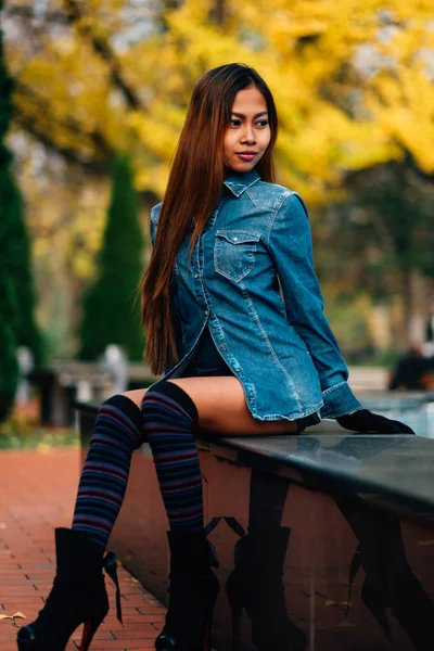 ファッショナブルな若い女性が高い丘と長いストライプ ソックス ブルー ジーンズのジャケットを身に着けています。公園で座っている女の子 — ストック写真