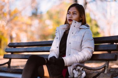  Genç güzel kadın şık sıcak kıyafeti bankta güneşli bir sonbahar günde parkta oturmuş portre. Şehir içinde rahat yaşam tarzı