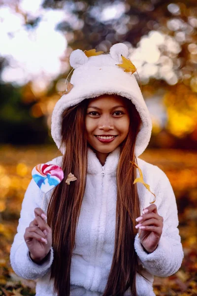 Young woman in the park on sunny autumn day, smiling, holding leaves and candy. Cheerful beautiful girl in white sweater in the park with nice colorful background