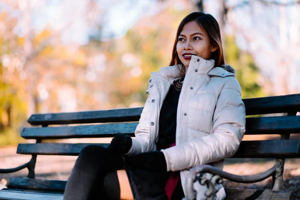  Genç güzel kadın şık sıcak kıyafeti bankta güneşli bir sonbahar günde parkta oturmuş portre. Şehir içinde rahat yaşam tarzı