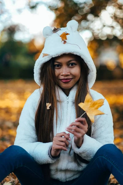  Sonbahar güneşli parkta genç kadın gülümseyerek, yaprakların tutmak. Beyaz kazaklı neşeli güzel kız