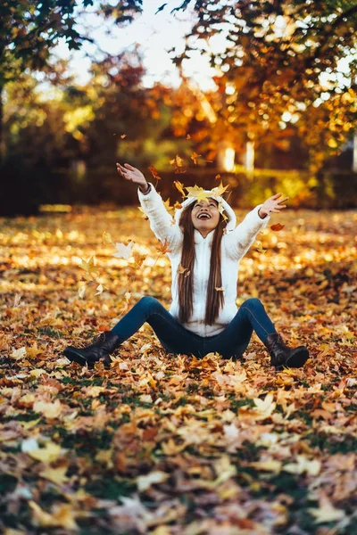 Happy young woman in park on sunny autumn day, laughing, playing leaves. Cheerful beautiful girl in white sweater during autumn season in the park