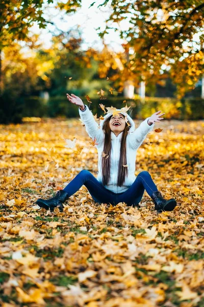 Gülüyor, yaprakları oynarken mutlu genç kadın sonbahar güneşli, parkta. Parkta sonbahar mevsiminde beyaz süveterli neşeli güzel kız