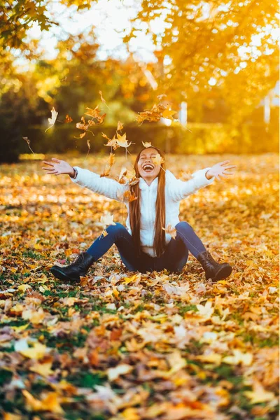 Happy young woman in park on sunny autumn day, laughing, playing leaves. Cheerful beautiful girl in white sweater during autumn season in the park