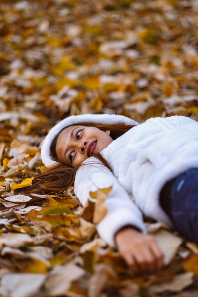 Autumn girl playing in city park. Portrait of an autumn woman lying over leaves and smiling outside in fall forest. Beautiful energetic Asian young woman