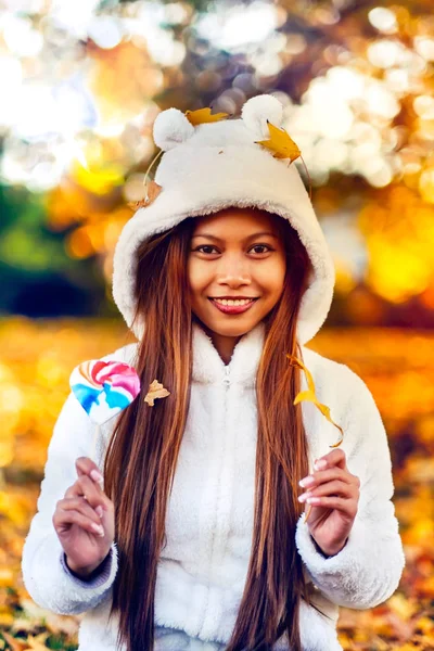 Young woman in the park on sunny autumn day, smiling, holding leaves and candy. Cheerful beautiful girl in white sweater in the park with nice colorful background