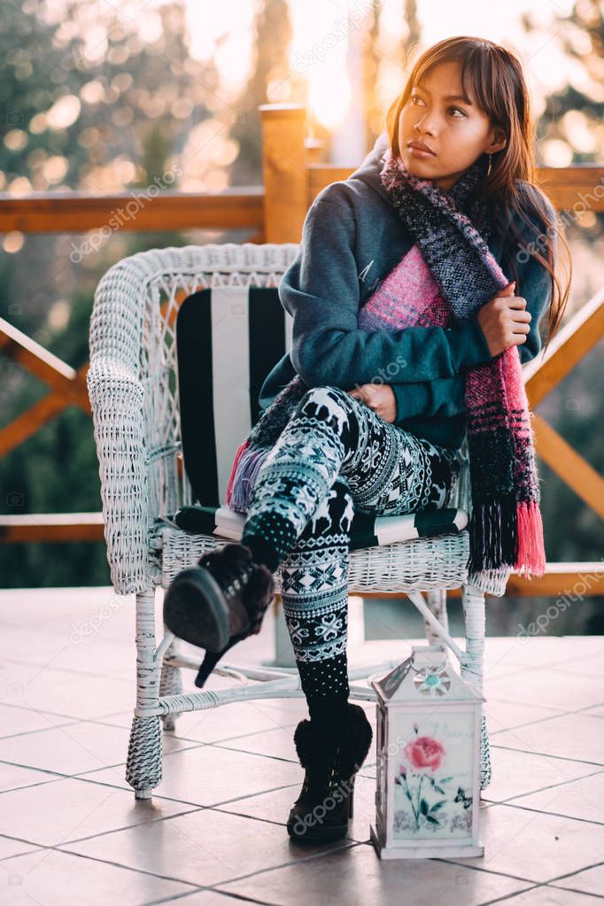 Young beautiful wman in a blue sweater with colorful scarf sitting on a white chair during cold weather outside the balcony