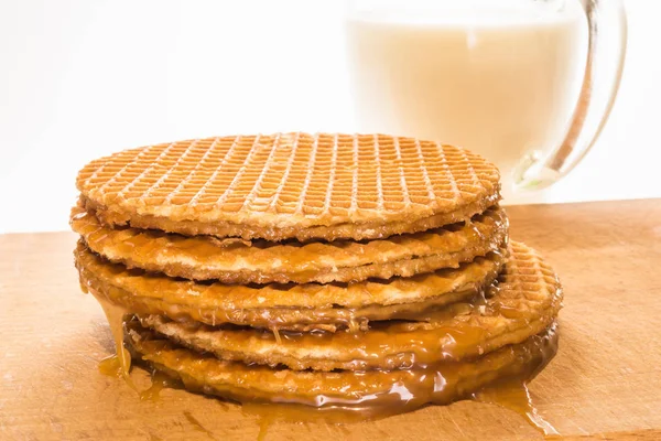 Galletas redondas u obleas con una capa de caramelo se encuentra en una pila áspera sobre una tabla de madera, taza de vidrio transparente con leche — Foto de Stock