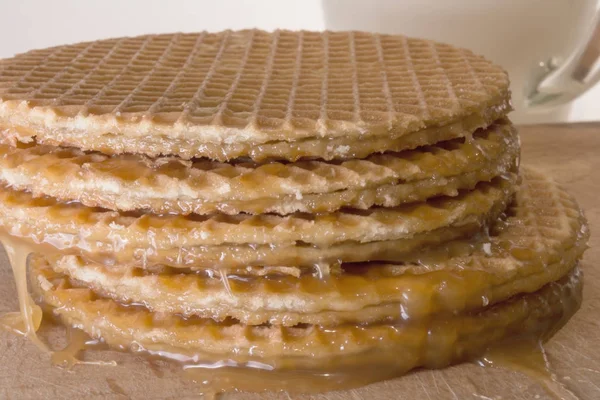 Galletas redondas u obleas con una capa de caramelo se encuentra en una pila áspera sobre una tabla de madera, taza de vidrio transparente con leche — Foto de Stock