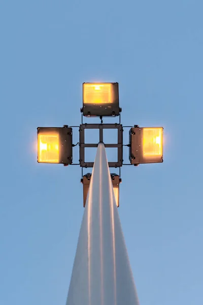 Street lamp with four floodlights shines with yellow light in the evening against a blue dark sky, perspective view from the bottom of a round post — Stock Photo, Image