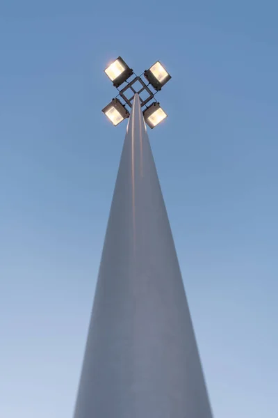 Lâmpada de rua com quatro holofotes brilha com luz amarela à noite contra um céu azul escuro, perspectiva vista do fundo de um poste redondo — Fotografia de Stock