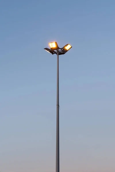 Street lamp with four floodlights shines with yellow light in the evening against a blue dark sky, perspective view from the bottom of a round post — Stock Photo, Image