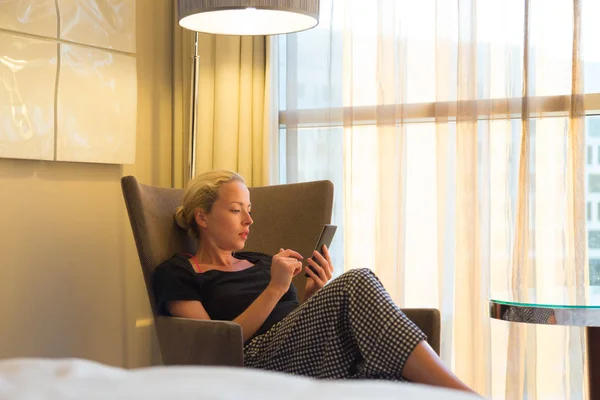 Atractiva mujer sosteniendo y utilizando el teléfono inteligente mientras se sienta en el sillón en la habitación de hotel moderna . — Foto de Stock