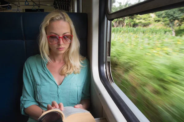 Blonde caucasian woman reading book on train by the window.