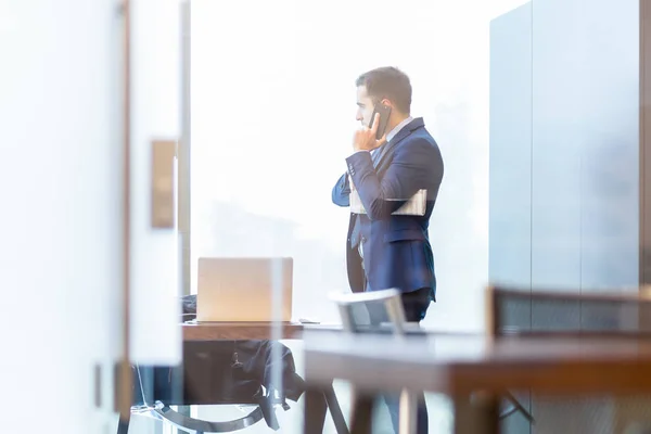 Zakenman praat op een mobiele telefoon terwijl hij door het raam kijkt. — Stockfoto