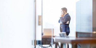 İş adamı pencereden bakarken cep telefonuyla konuşuyor.