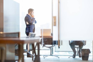 İş adamı pencereden bakarken cep telefonuyla konuşuyor.