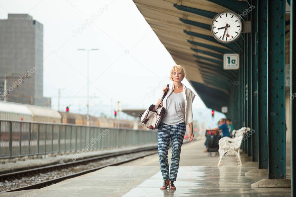 Woman waiting at platform of railway station bearing vintage shoulder bag.