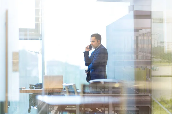 Empresario hablando en un teléfono móvil mientras mira por la ventana de la oficina . —  Fotos de Stock