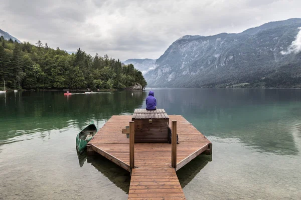 Lila kapucnis nő nyugodt, borús reggeli jelenetet néz a Bohinj tónál, Alpokban, Szlovéniában. — Stock Fotó