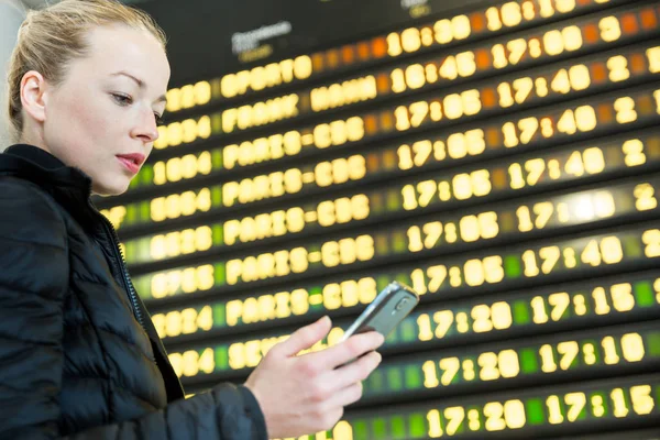 Mulher no aeroporto em frente à placa de informações de voo verificando seu telefone . — Fotografia de Stock