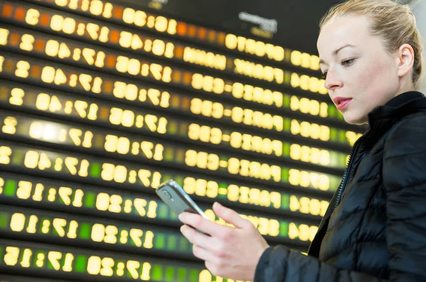 Mulher no aeroporto em frente à placa de informações de voo verificando seu telefone . — Fotografia de Stock