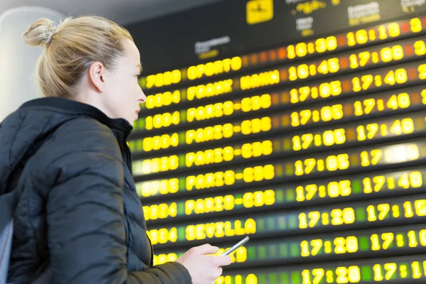 Mujer en el aeropuerto delante del tablero de información de vuelo revisando su teléfono . —  Fotos de Stock