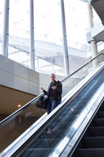 Bizneswoman z duża czarna torba i telefon komórkowy malejącej na ruchomych schodach. — Zdjęcie stockowe