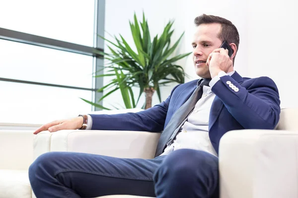 Bajo ángulo de tiro de un joven hombre de negocios guapo en un elegante espacio de oficina moderno con grandes ventanas, hablando por teléfono móvil . — Foto de Stock