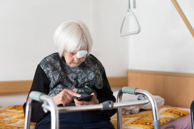Lonley elderly 95 years old woman sitting at the bad using modern mobile phone. clipart