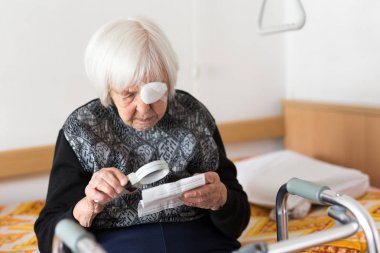 Visually impaired elderly 95 years old woman sitting at the bad trying to read with magnifying glass. clipart