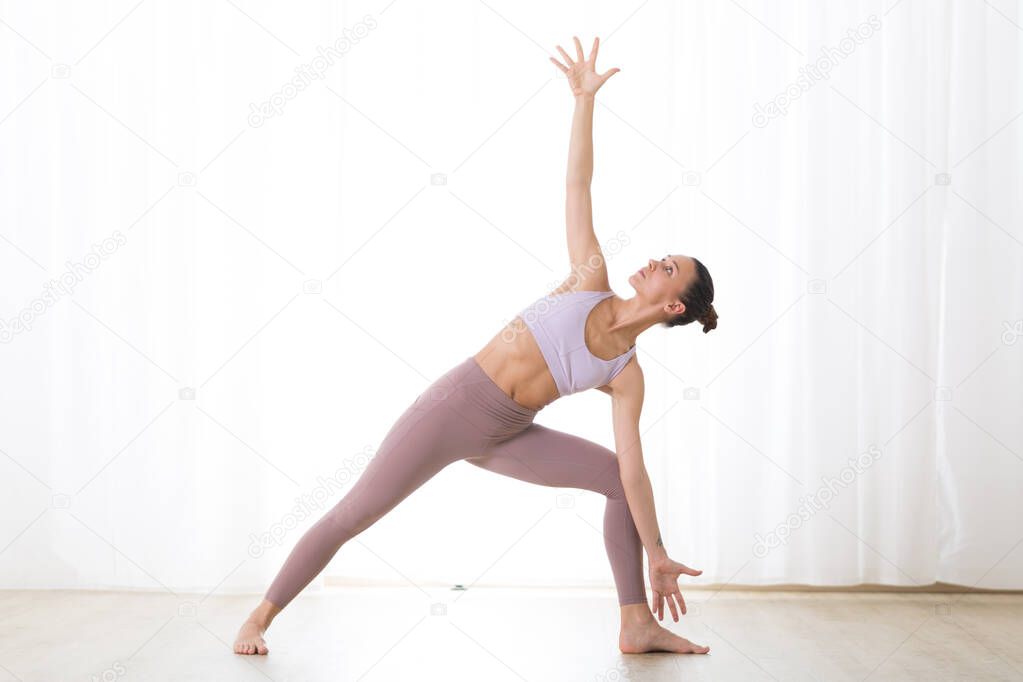 Portrait of gorgeous active sporty young woman practicing yoga in studio. Beautiful girl practice Trikonasana, triangle yoga pose. Healthy active lifestyle, working out indoors in gym