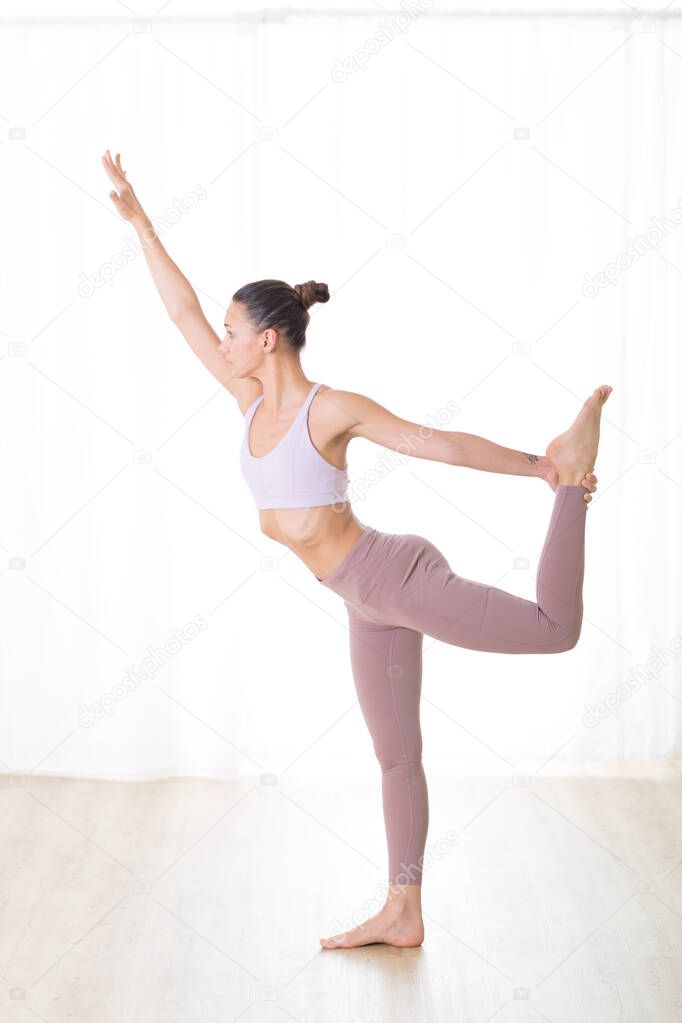 Portrait of gorgeous active sporty young woman practicing yoga in studio. Beautiful girl practice Dandayamana Dhanurasana, standing bow pulling pose. Healthy active lifestyle, working out in gym