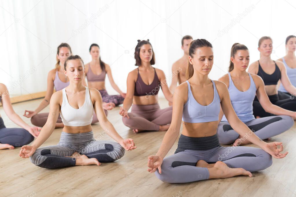 Group of young sporty attractive people in yoga studio, practicing yoga lesson with instructor, sitting on floor in Siddhasana, easy seated yoga pose. Healthy active lifestyle, working out in gym