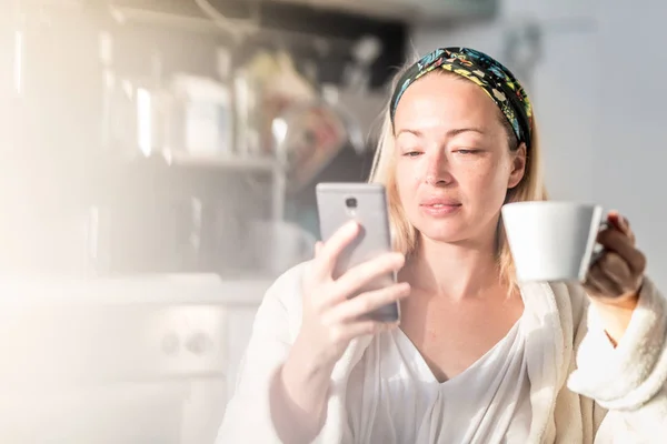 Schöne kaukasische Frau zu Hause, fühlt sich wohl im weißen Bademantel, nimmt sich Zeit für sich, trinkt Morgenkaffee und liest morgens Nachrichten auf dem Handy — Stockfoto