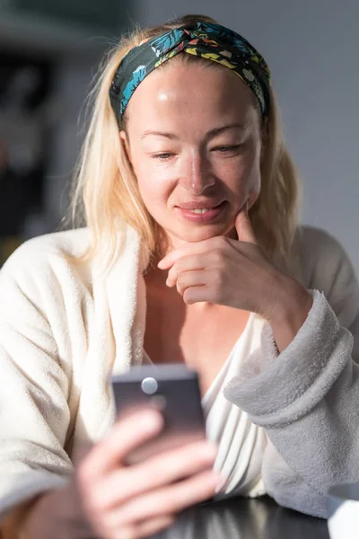 Vacker kaukasiska kvinna hemma, känna sig bekväm klädd i vit badrock, ta lite tid för sig själv, dricka morgonkaffe och läsa nyheter på mobiltelefon enhet på morgonen — Stockfoto