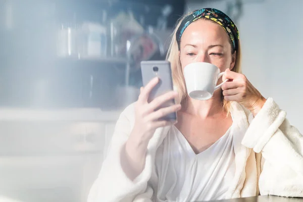 Schöne kaukasische Frau zu Hause, fühlt sich wohl im weißen Bademantel, nimmt sich Zeit für sich, trinkt Morgenkaffee und liest morgens Nachrichten auf dem Handy — Stockfoto