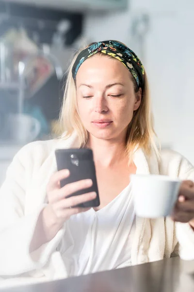 Piękna biała kobieta w domu, czuje się komfortowo nosząc biały szlafrok, spędzając trochę czasu dla siebie, pijąc poranną kawę i czytając wiadomości na telefonie komórkowym rano — Zdjęcie stockowe