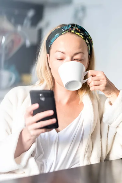 Piękna biała kobieta w domu, czuje się komfortowo nosząc biały szlafrok, spędzając trochę czasu dla siebie, pijąc poranną kawę i czytając wiadomości na telefonie komórkowym rano — Zdjęcie stockowe