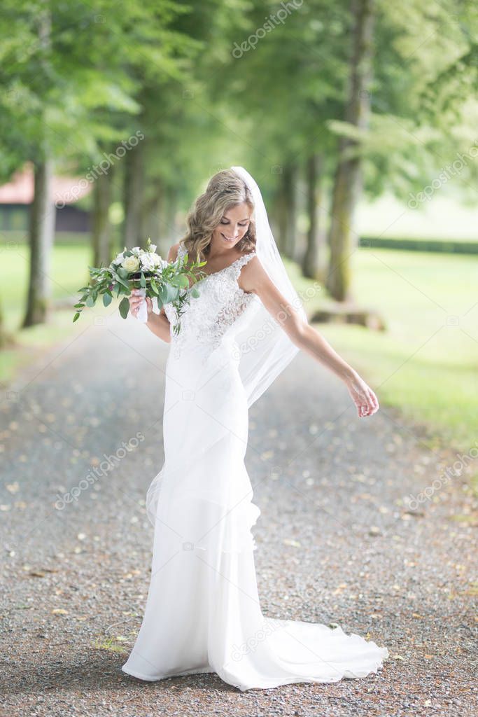 Full length portrait of beautiful sensual young blond bride in long white wedding dress and veil, holding bouquet outdoors in natural background