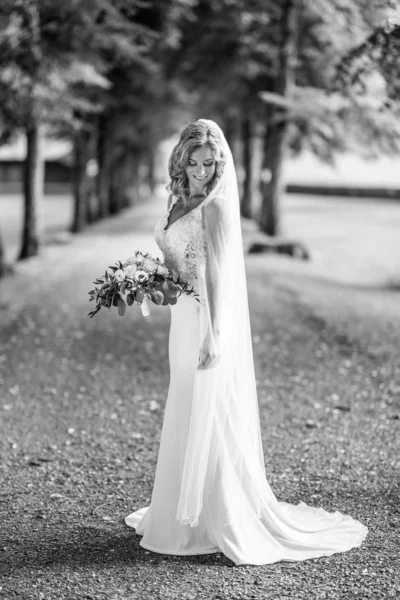 長い白いウェディングドレスとベールで美しい官能的な若いブロンドの花嫁の完全な長さの肖像画、自然な背景で屋外で花束を保持. — ストック写真