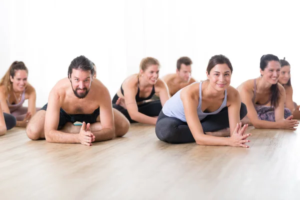 Group of young sporty attractive people in yoga studio, practicing yoga lesson with instructor, sitting on floor in forward band stretching yoga pose. Healthy active lifestyle, working out in gym — Stock Photo, Image