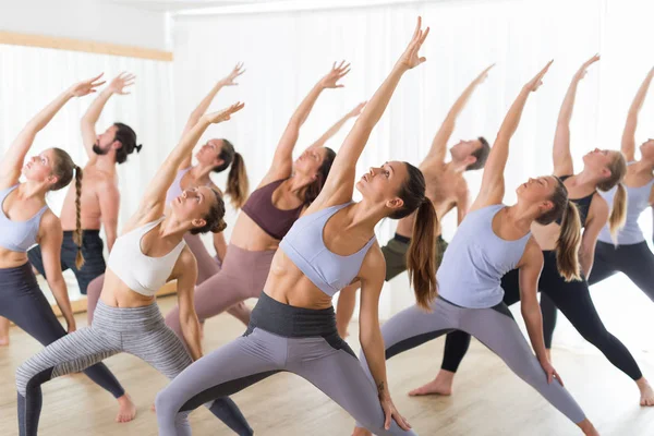 Groep van jonge sportieve aantrekkelijke mensen in yoga studio, het beoefenen van yoga les met instructeur, stretching op de vloer in Trikonosana, driehoek yoga pose. Gezonde actieve levensstijl, sporten in de sportschool. — Stockfoto