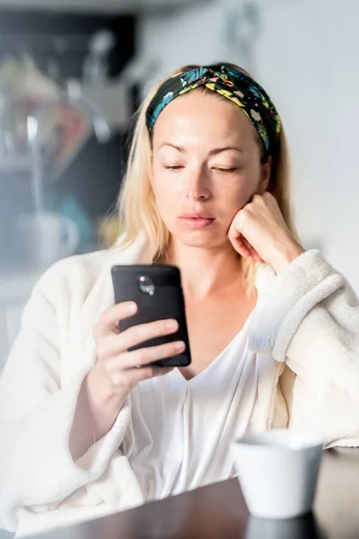 Hermosa mujer caucásica en casa, sintiéndose cómoda usando albornoz blanco, tomando algún tiempo para sí misma, bebiendo café de la mañana y leyendo noticias en el dispositivo de teléfono móvil por la mañana — Foto de Stock