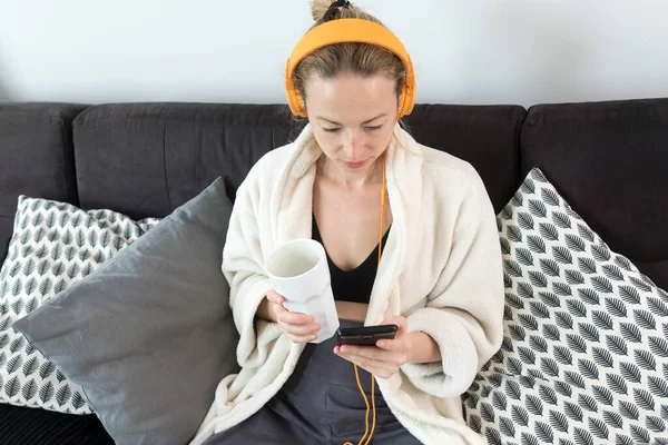 Reste à la maison. Distance sociale. Femme à la maison relaxant sur canapé canapé boire du thé de tasse blanche, écouter de la musique relaxante, rester connecté aux amis et à la famille via les réseaux sociaux sur mobile . — Photo