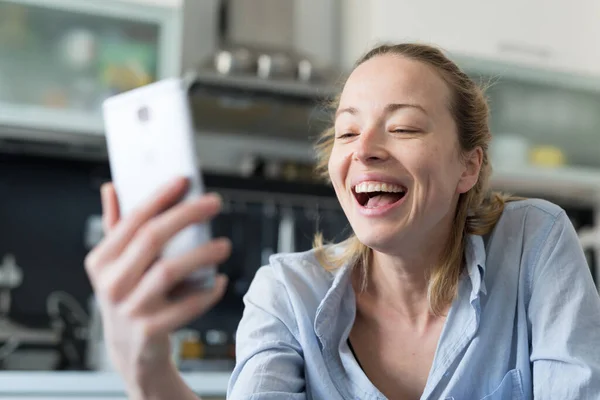 Unga leende glad nöjd kvinna inomhus i köket med hjälp av sociala medier apps på mobiltelefon för att chatta och sticka i samband med sina nära och kära. Stanna hemma, social distanserande livsstil. — Stockfoto