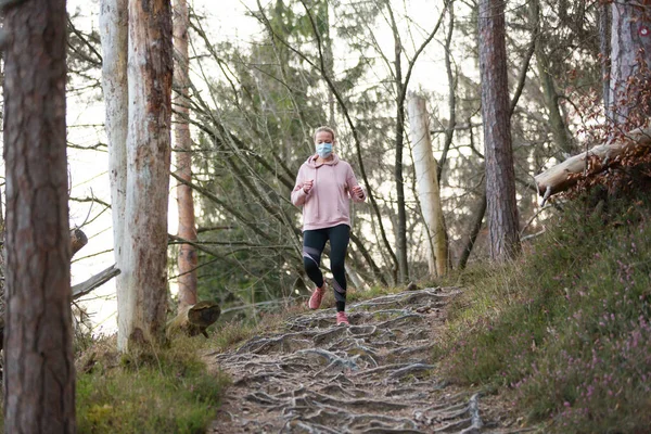 Corona virus, or Covid-19, is spreading all over the world. Portrait of caucasian sporty woman wearing a medical protection face mask while running in nature. — Stock Photo, Image